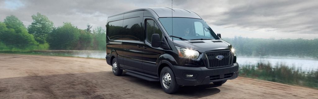 Black Ford Transit van driving on a road near a lake.