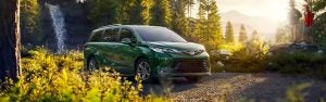 A green Toyota Sienna parked on the edge of a forest, surrounded by yellow flower.