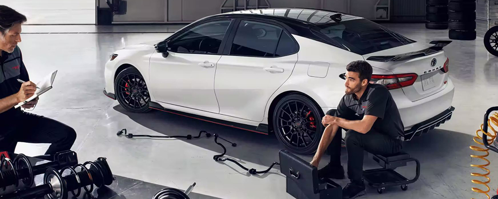 Two mechanics inspect a white, Toyota Sedan.