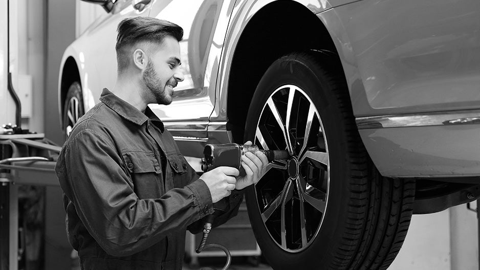 A grayscale image of a smiling mechanic using an impact wrench to change a tire on a vehicle lifted on a service rack.