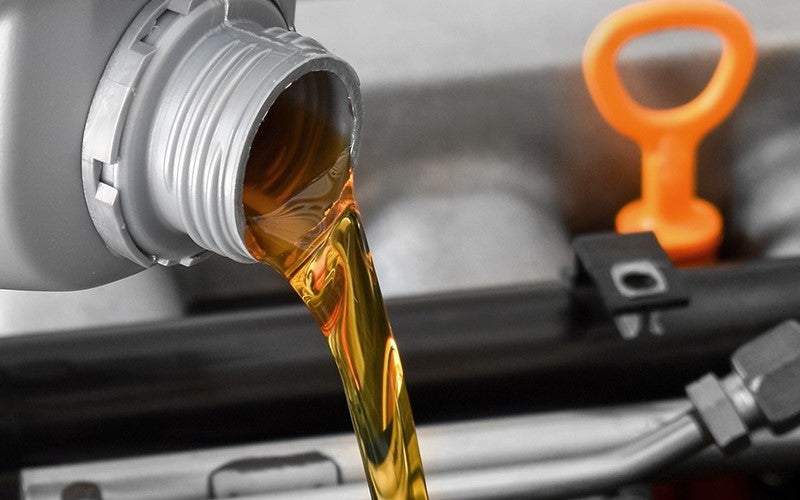 Close-up of fresh, amber-colored engine oil being poured into a vehicle's engine from a silver bottle.