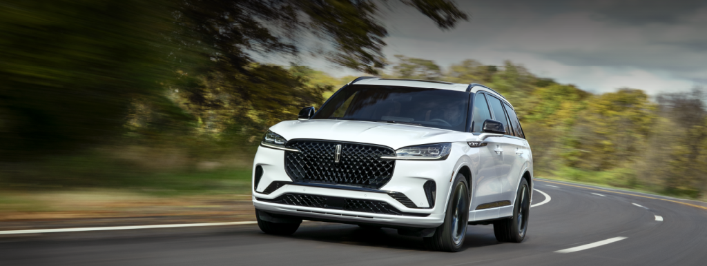 A white 2025 Lincoln Aviator driving along a highway bordered by a forest of oak trees near London, KY.