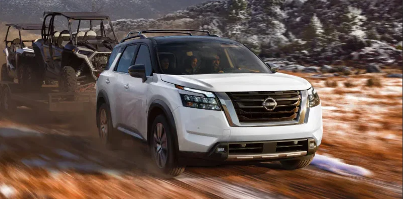 A white Nissan Pathfinder driving down a dirt road towing ATVs behind it.