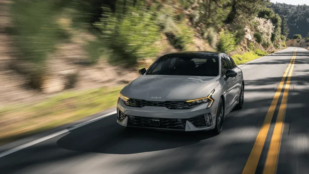 Front view of 2026 Kia K5 driving through scenic highway with sporty and aerodynamic styling.
