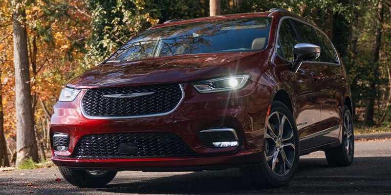 Front corner view of a red Chrysler Pacifica minivan, headlights glowing, parked on an asphalt road 