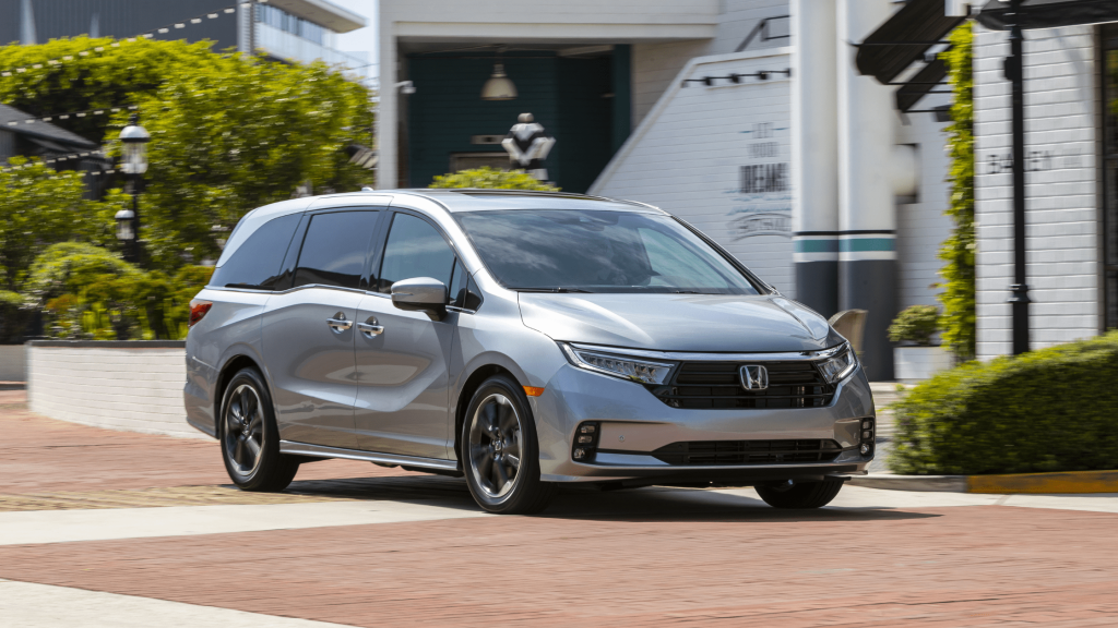 A silver 2024 Honda Odyssey minivan driving through a modern urban area with green landscaping and stylish architecture in the background.