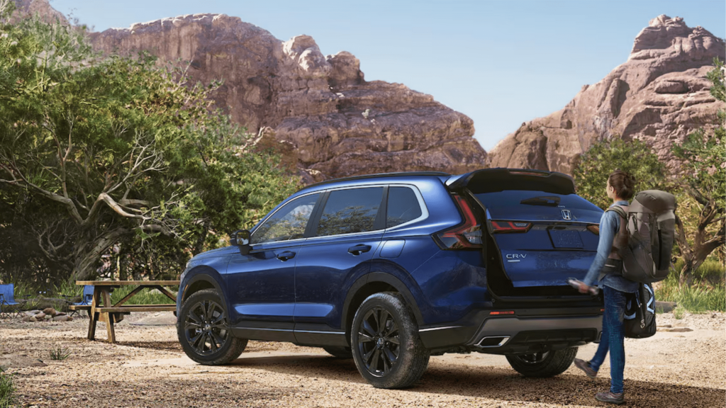 A person with a backpack stands next to a blue 2024 Honda CR-V Hybrid SUV in a scenic outdoor setting with rocky mountains and trees in the background.