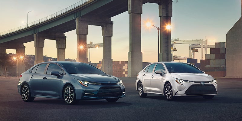 Two cars, one blue and one white, are parked under a large overpass at dusk. The setting sun casts a warm glow, creating an urban, modern atmosphere.