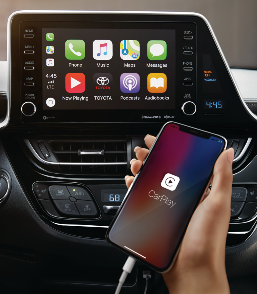 Close-up of a car dashboard displaying a touchscreen with Apple CarPlay apps. A hand holds a connected phone showing the CarPlay logo, conveying modern technology.