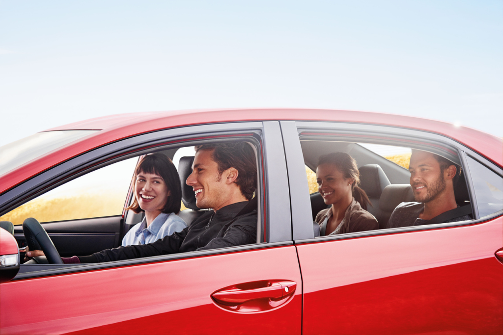 Four people are smiling and chatting inside a moving red car with an open window. The sky is clear, suggesting a pleasant, sunny day.