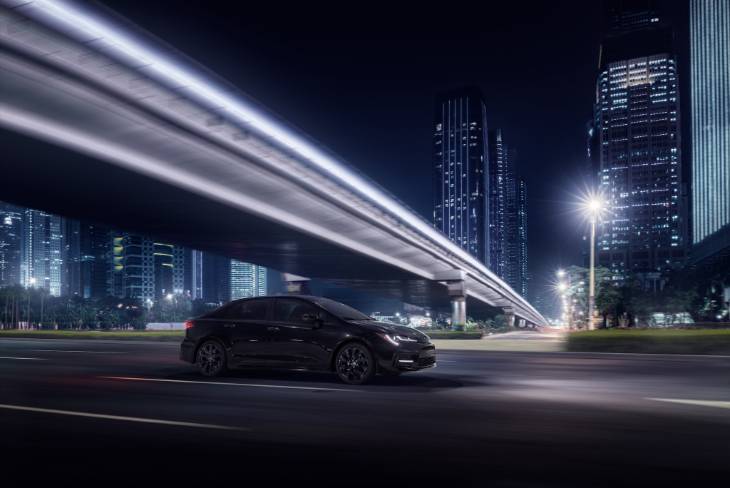Sleek black car driving on a city road at night, with illuminated skyscrapers and a modern bridge overhead. The scene is dynamic and futuristic.