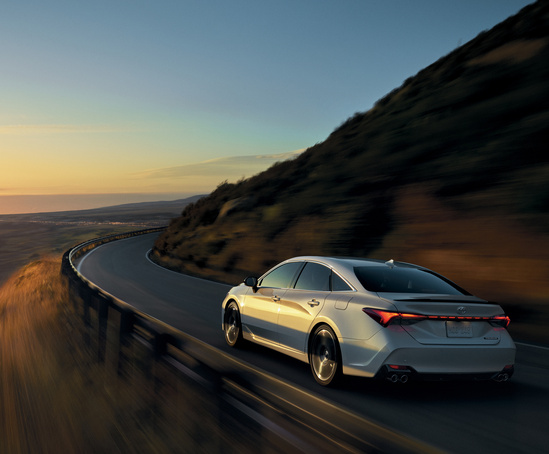 A sleek white car drives along a curvy mountain road at sunset, conveying freedom and adventure. The sky is warm with orange hues, casting soft light.