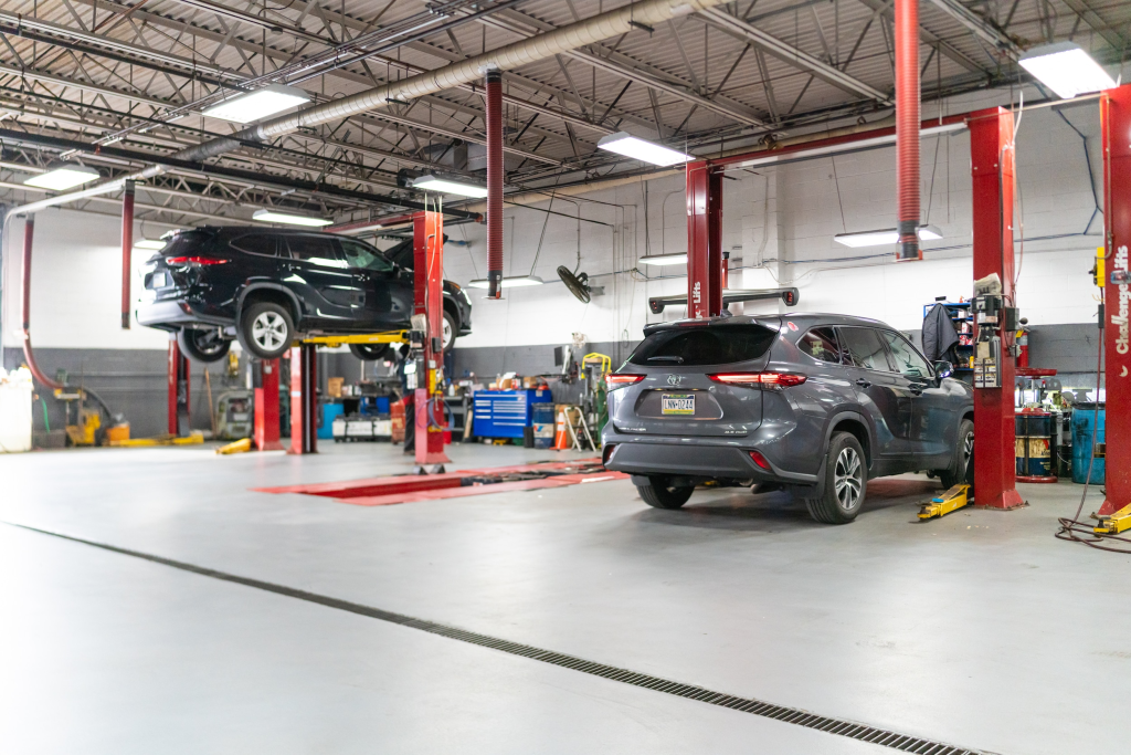 A spacious auto repair shop with a car lifted on a red hydraulic lift and another vehicle parked nearby. Bright overhead lighting and organized tools create a professional atmosphere.