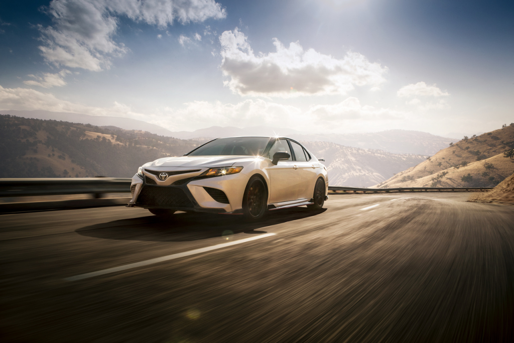 A white car cruises along a winding mountain road under a clear blue sky. Sunlight reflects off the hood, suggesting speed and freedom.