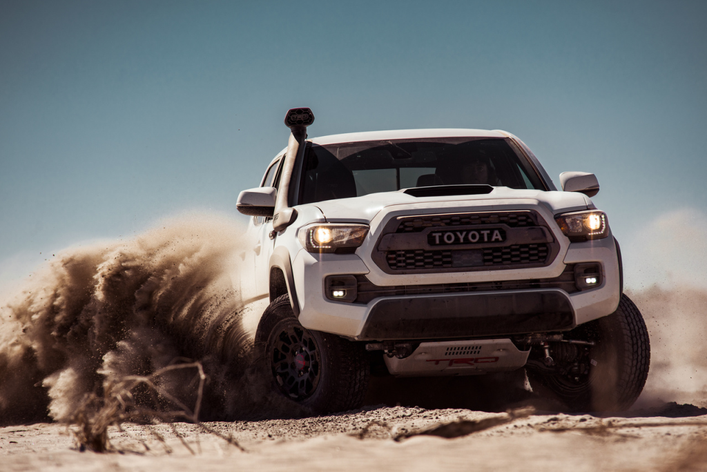 White Toyota truck driving through desert sand, creating a dramatic dust cloud. The scene conveys action, adventure, and rugged power under a clear sky.