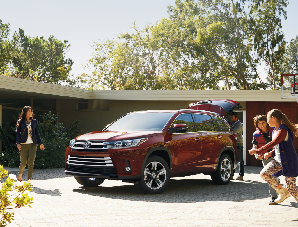 Family gathers around a shiny red SUV with an open trunk, parked in a sunlit driveway. Trees and a modern house in the background evoke a joyful, carefree atmosphere.