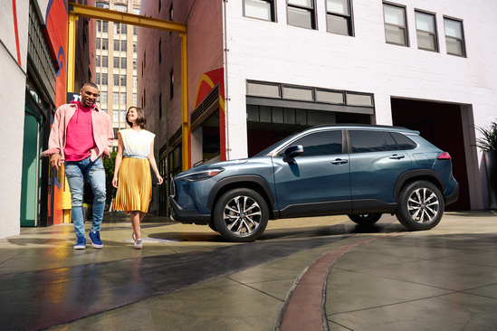 A smiling couple walks down a colorful alley beside a parked blue SUV. The setting is urban and vibrant, conveying a sense of joy and exploration.