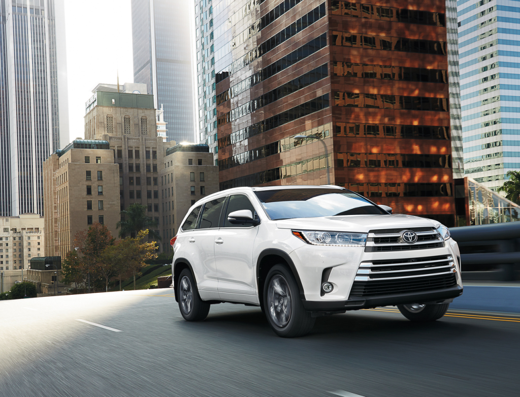 A white SUV drives on a city street flanked by tall buildings reflecting sunlight. The scene conveys urban sophistication and dynamic motion.