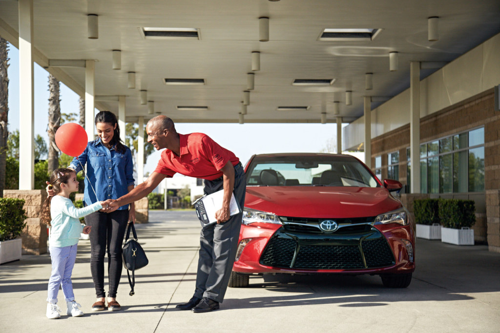 A smiling salesman in a red shirt shakes hands with a young girl holding a red balloon, next to her mother. A red Toyota car is parked behind them.
