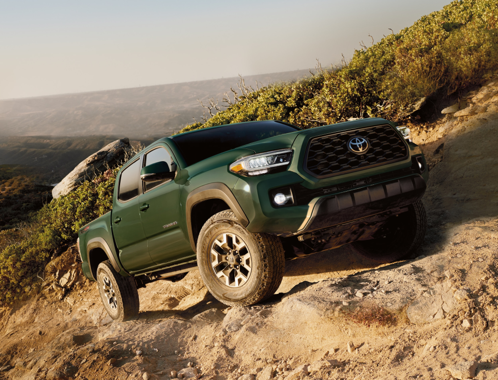 A green pickup truck ascends a rugged, sandy trail surrounded by sparse greenery, showcasing its off-road capabilities under a clear, expansive sky.