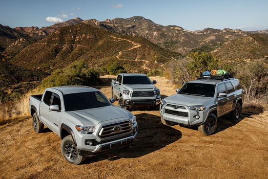Three silver trucks are parked on a dirt path against a backdrop of rugged mountains. The vehicles convey a sense of adventure and exploration.