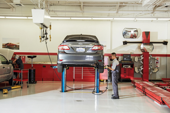 A mechanic in uniform checks a gray car lifted on a blue hydraulic lift in a tidy auto repair shop with red tool cabinets and clean white floors.