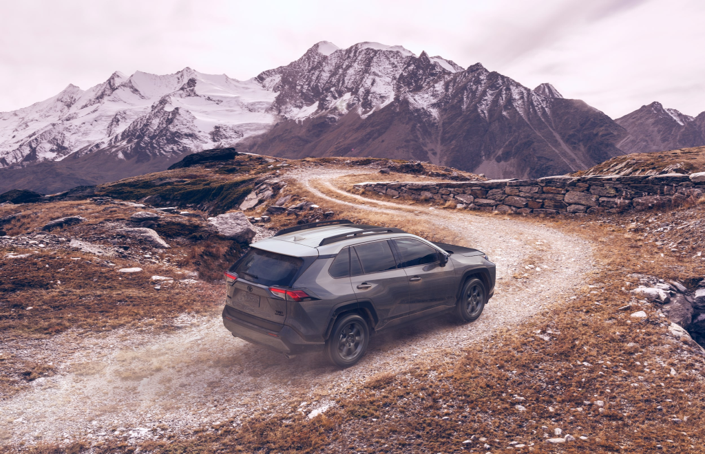 SUV driving on a winding gravel road in a mountainous landscape with snow-capped peaks. The scene feels adventurous and serene.