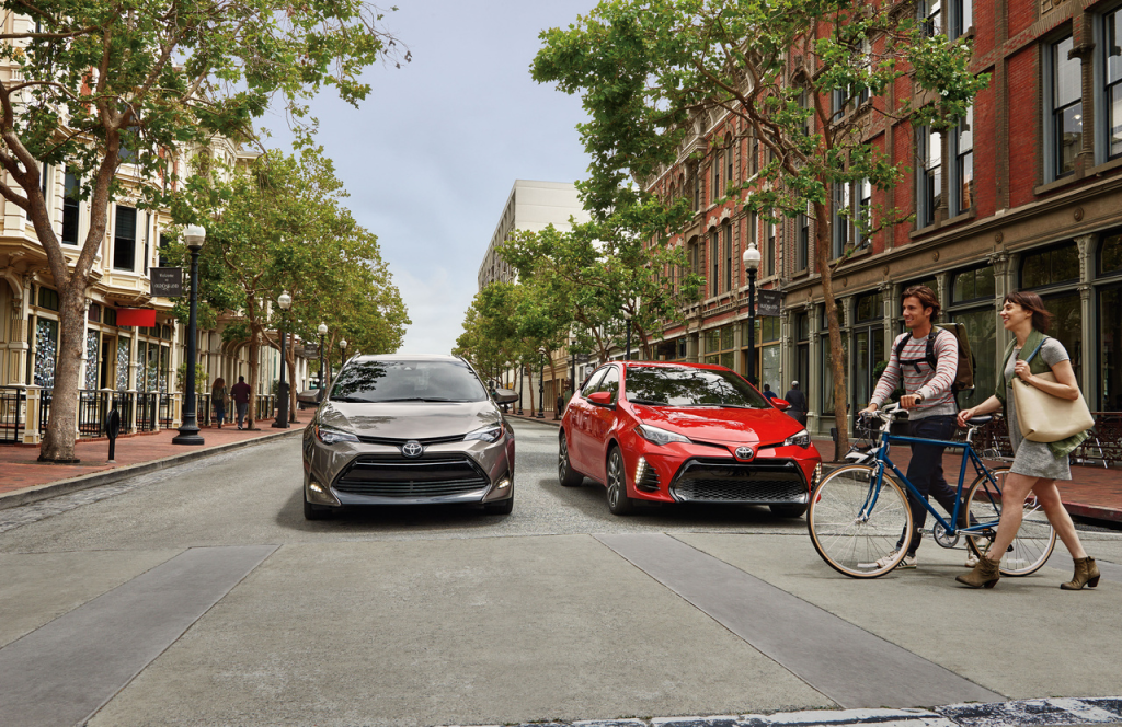 Two Toyota cars, one gray and one red, are parked on a tree-lined street. A couple walks with a bicycle, conveying a lively and urban atmosphere.