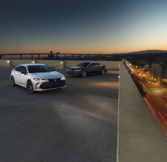 Two sedans, one white and one dark, are parked on a rooftop at dusk. City lights and traffic trails are visible, creating a modern, serene vibe.