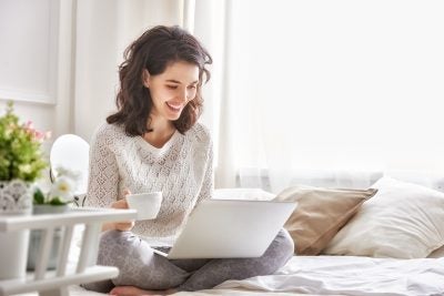 Woman sitting on a bed holding a cup of coffee on a laptop