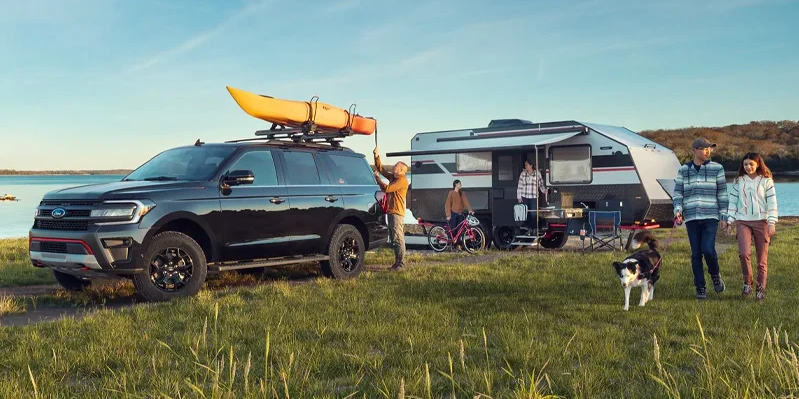 A photo of a 2025 Ford Expedition parked in a field with a kayak on the roof and a camper parked behind it.