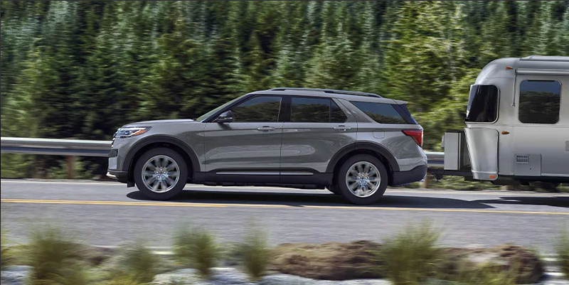 A Silver 2025 Ford Explorer towing a camper down the road.