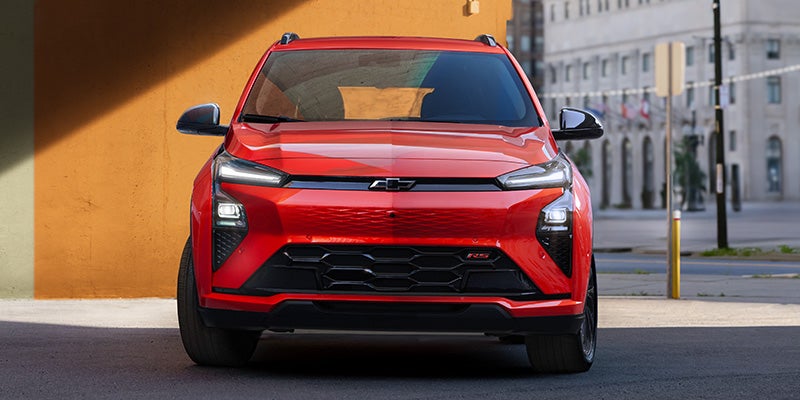 Front view of a bright red Chevrolet Bolt parked outdoors in front of a warm orange wall and city street background.