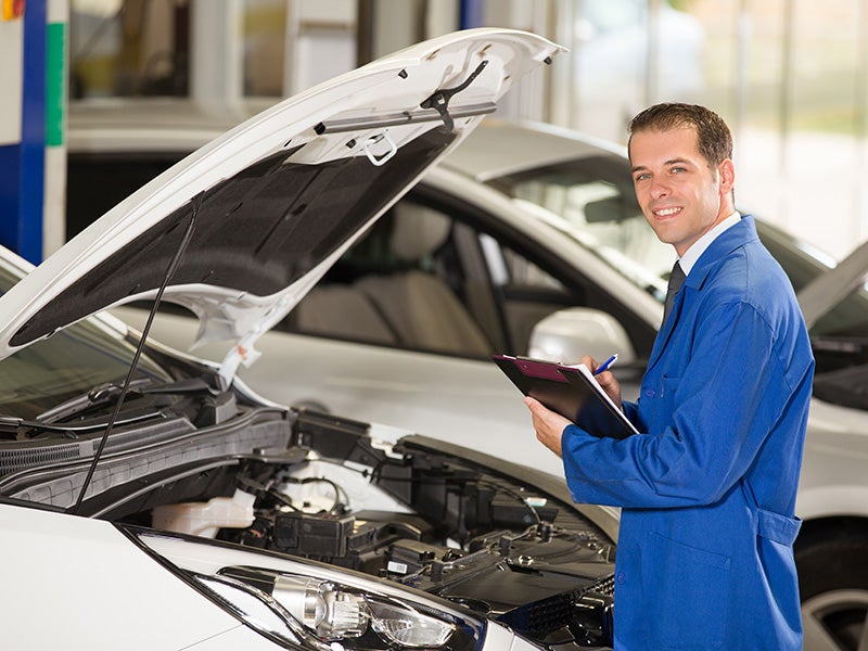 Smiling mechanic holding a clipboard next to an open car hood.