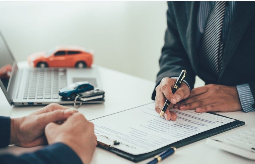 Financing process scene showing a person signing a document on a clipboard with a pen while discussing terms with another individual, with small toy cars and a laptop in the background suggesting an automotive sales or financing agreement.