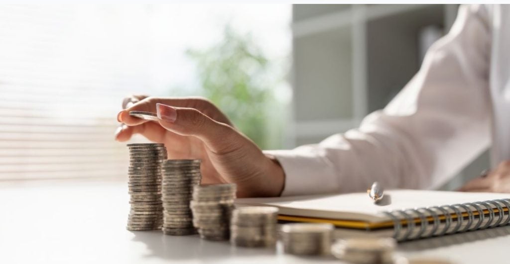 Hand reaching to stack coins on a desk, symbolizing savings or financial planning, with a notebook and pen in the background under soft natural light.