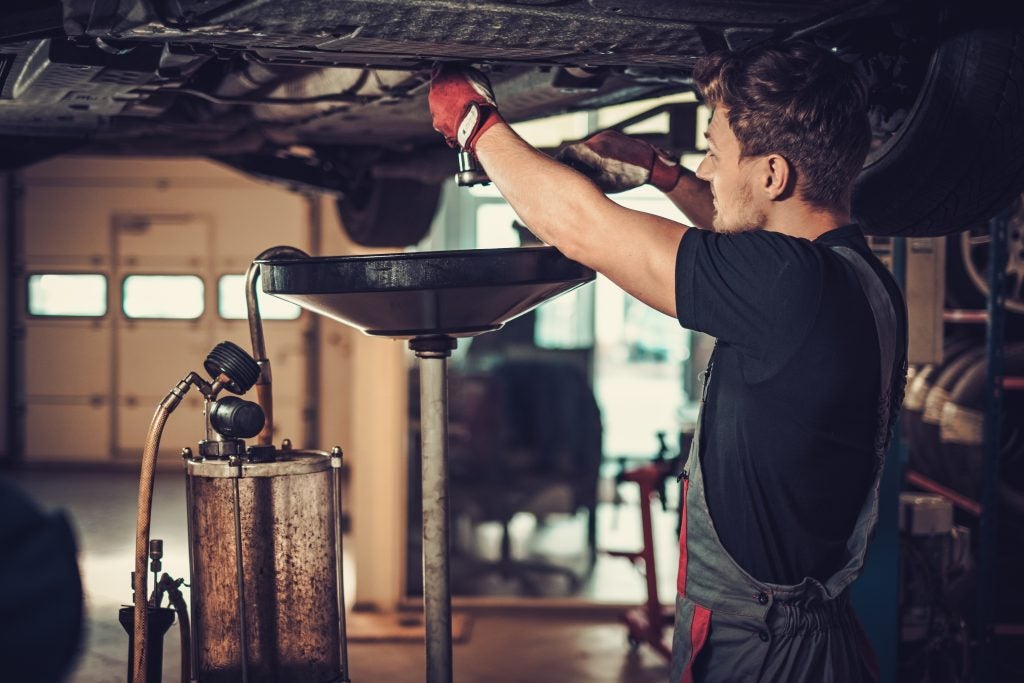 Mechanic working underneath a raised vehicle in an auto repair shop, performing an oil change using a drain pan while wearing protective gloves.