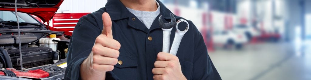 A mechanic giving a thumbs up while holding two wrenches, with a car engine in the background and a blurred workshop setting.