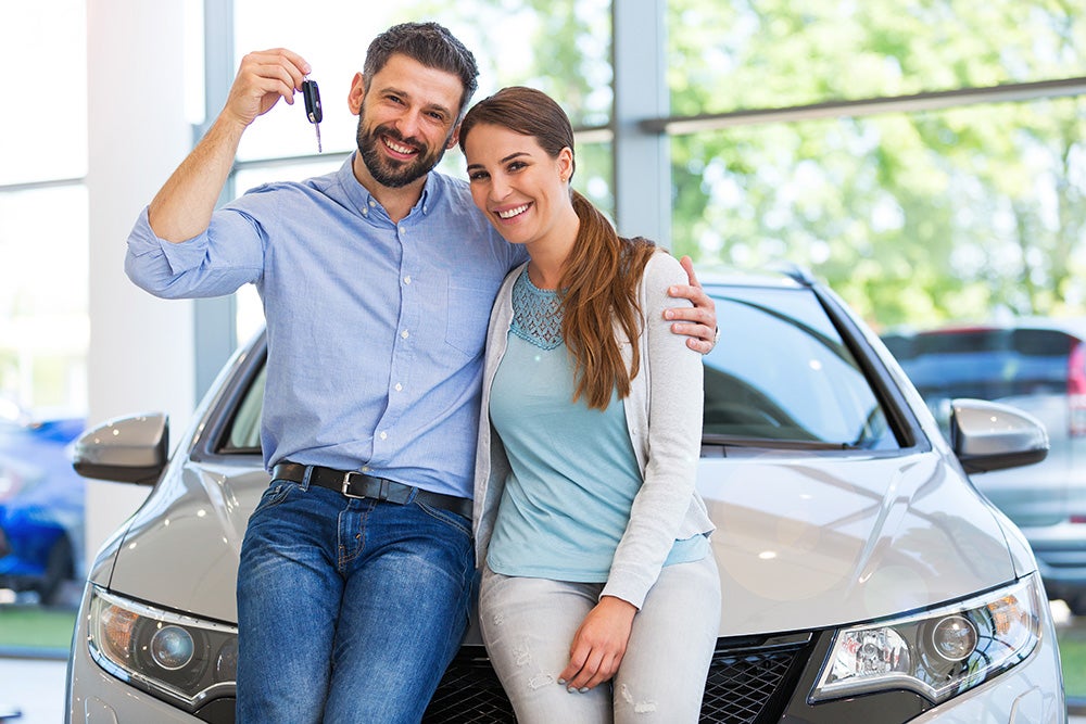 A smiling couple, the man holding up a car key, lean against a new car in a dealership showroom.