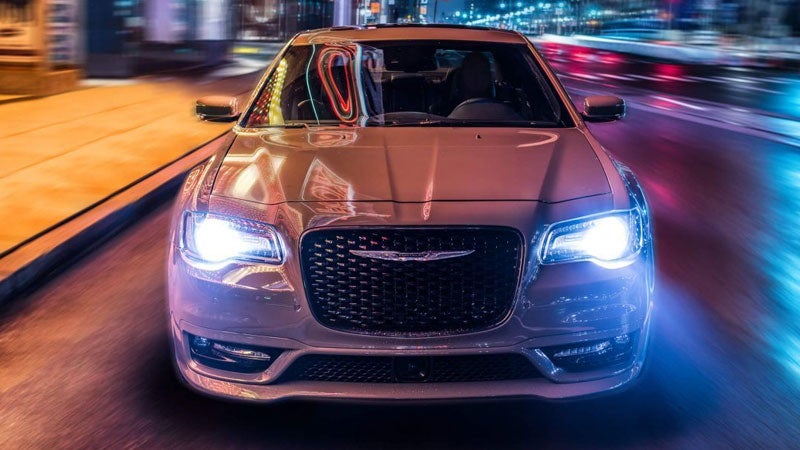 A silver car with bright headlights speeds down a colorful city street at night, creating light streaks.