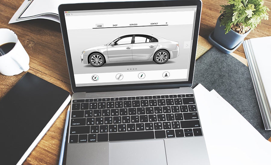 Laptop displaying a car sales website with a silver sedan, coffee mug, and plant on a wooden desk.