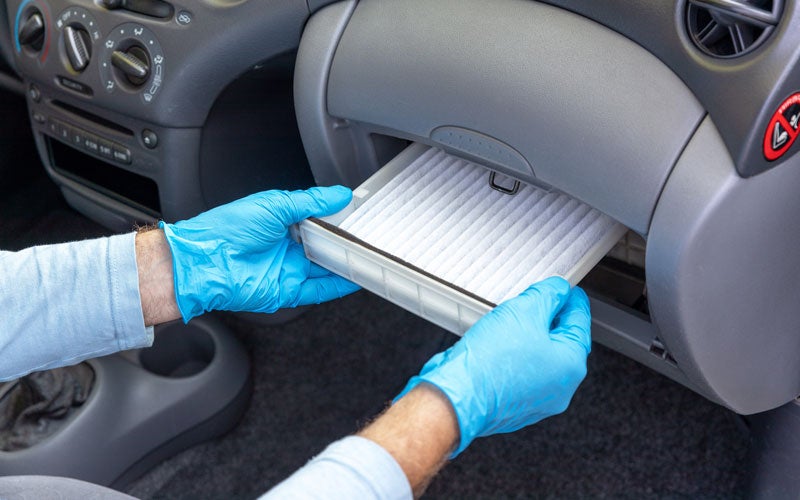 A person wearing blue gloves pulls out a white, rectangular cabin air filter from a car's open glove compartment.