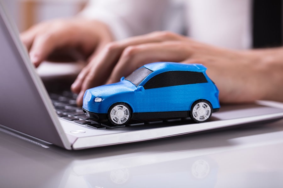 A blue toy car rests on a laptop keyboard. Hands are typing on the keyboard behind the car.