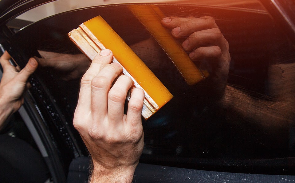 A person’s hands applying tint film to a car window with a squeegee, smoothing out bubbles.