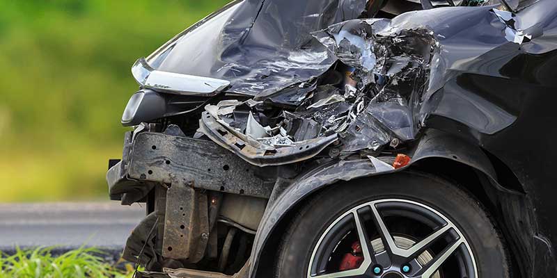 The front passenger side of a black car is severely damaged with the bumper detached and the hood crumpled upwards.