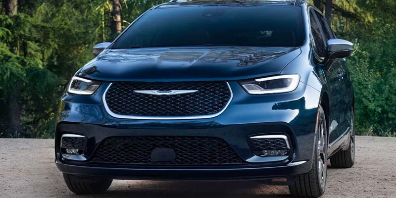 Front view of a dark blue Chrysler minivan parked outdoors, with a forest of green trees in the background.