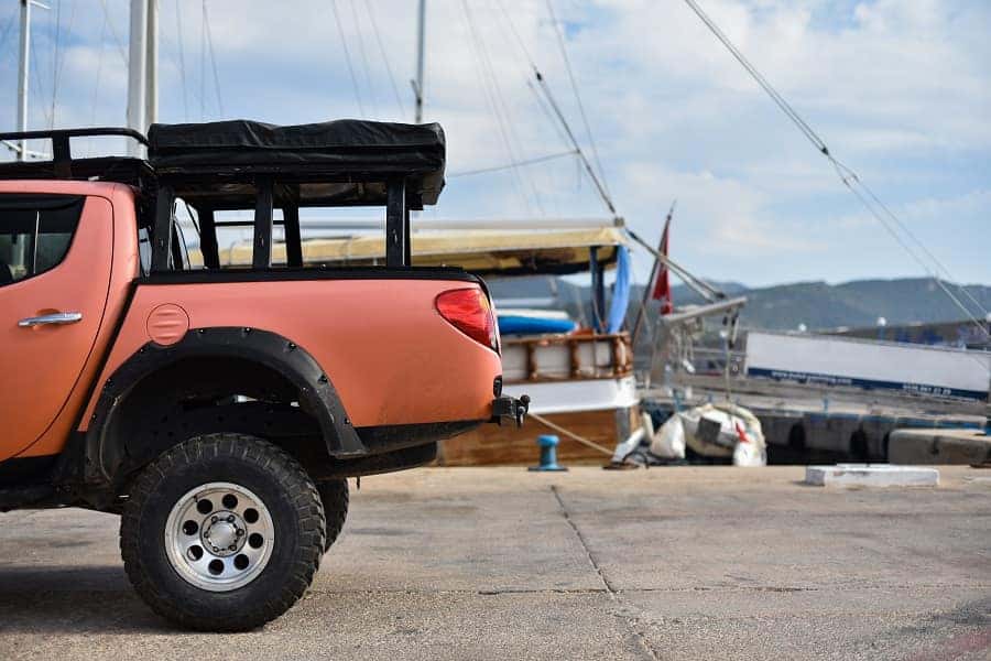 A coral-colored pickup truck with a black roof tent and roll bars, parked in a marina with boats.