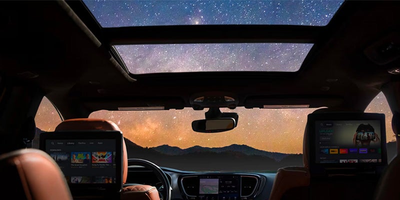 Interior view of a car with two screens on the back of the front seats showing multimedia content. The sunroof reveals a star-filled night sky over mountains.
