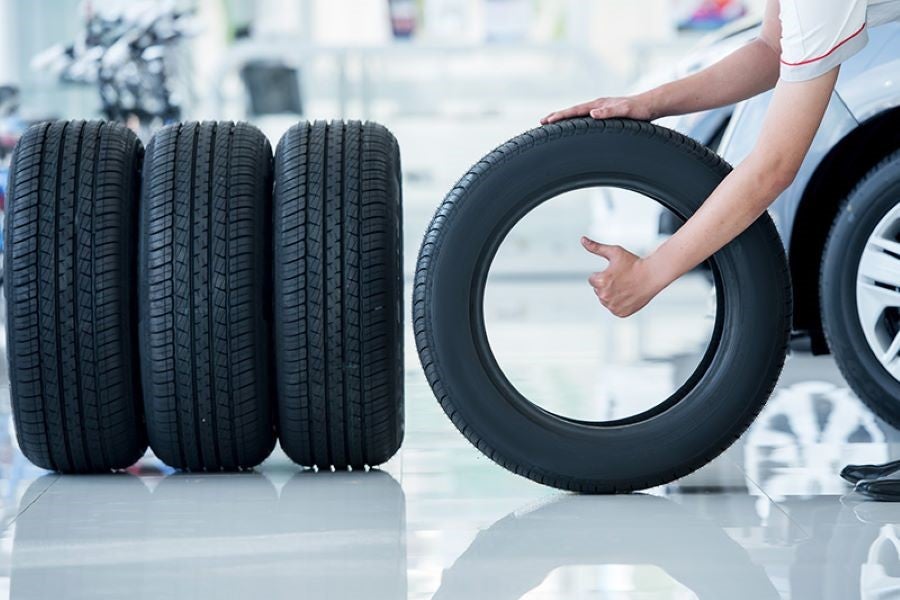 A person holds a new car tire upright, giving a thumbs up. Three other new tires stand next to it.