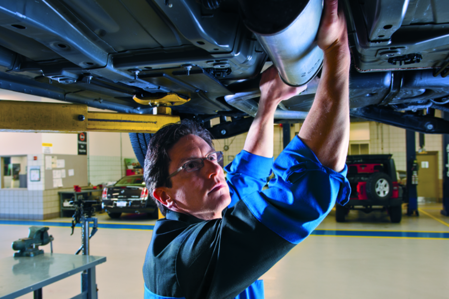 A male mechanic in glasses and a blue uniform works on the exhaust pipe of a car on a lift.